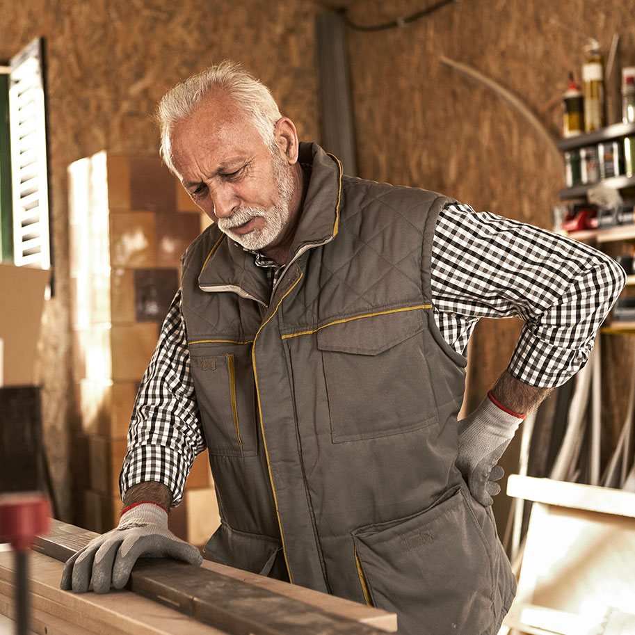 A man working in a wood shop hunches forward with his hand on his back. Workers’ compensation for spinal injuries covers many types of back trauma that you can suffer on the job.