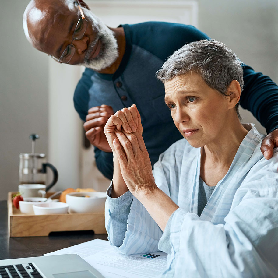 A woman sits at a laptop with a man standing and putting his hand on her shoulder. HNB can help you determine if you can speed up your cancer disability benefits.