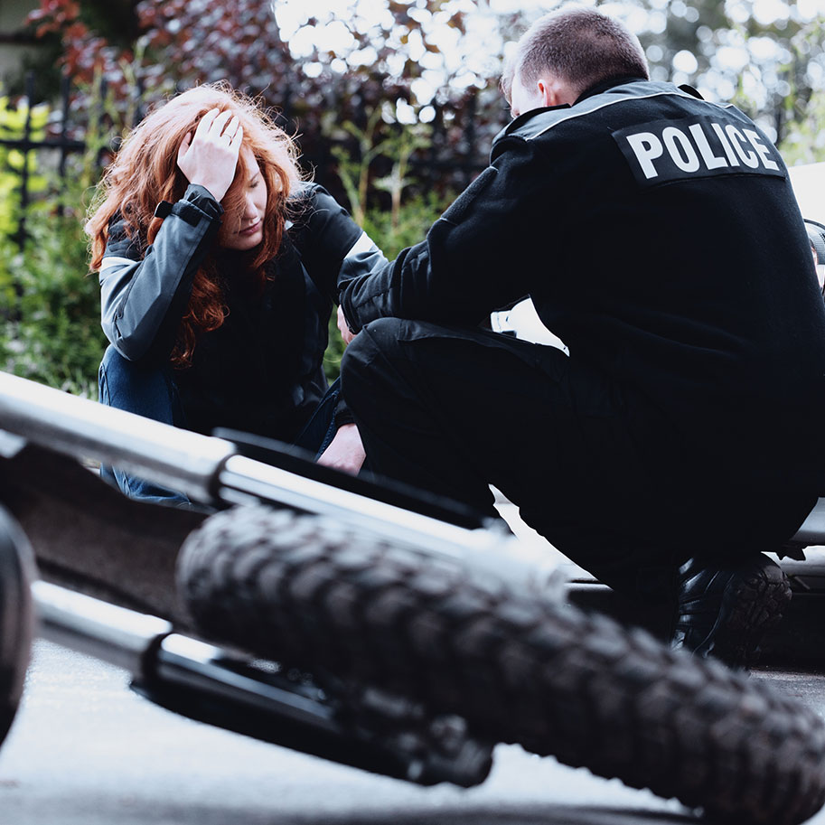 A motorcycle lies in the road while a police officer talks to a woman sitting on the curb. Unlike insurance companies, Horenstein, Nicholson & Blumenthal puts your rights as a motorcycle rider first after an accident.
